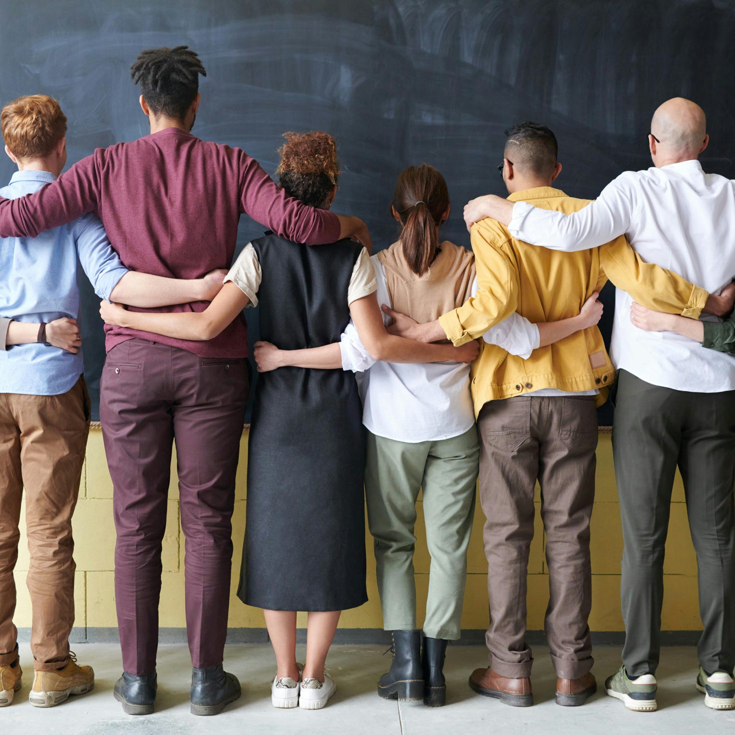 A diverse group of adults in casual outfits hugging in front of a chalkboard, symbolizing teamwork.