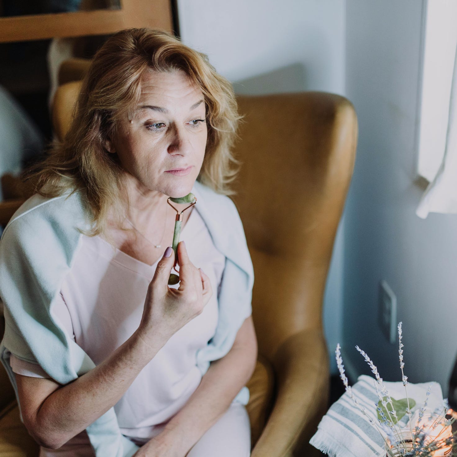Mature woman engaging in skincare ritual with facial roller indoors for self-care.